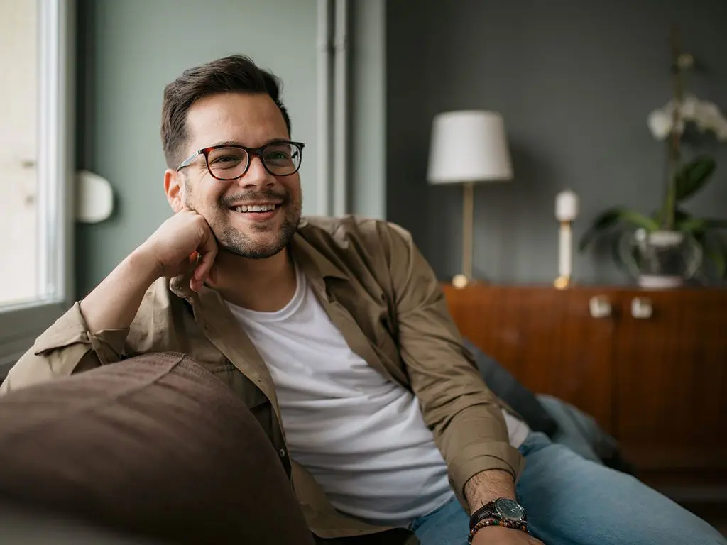 A sitting man in glasses leans comfortable on a brown sofa, smiling