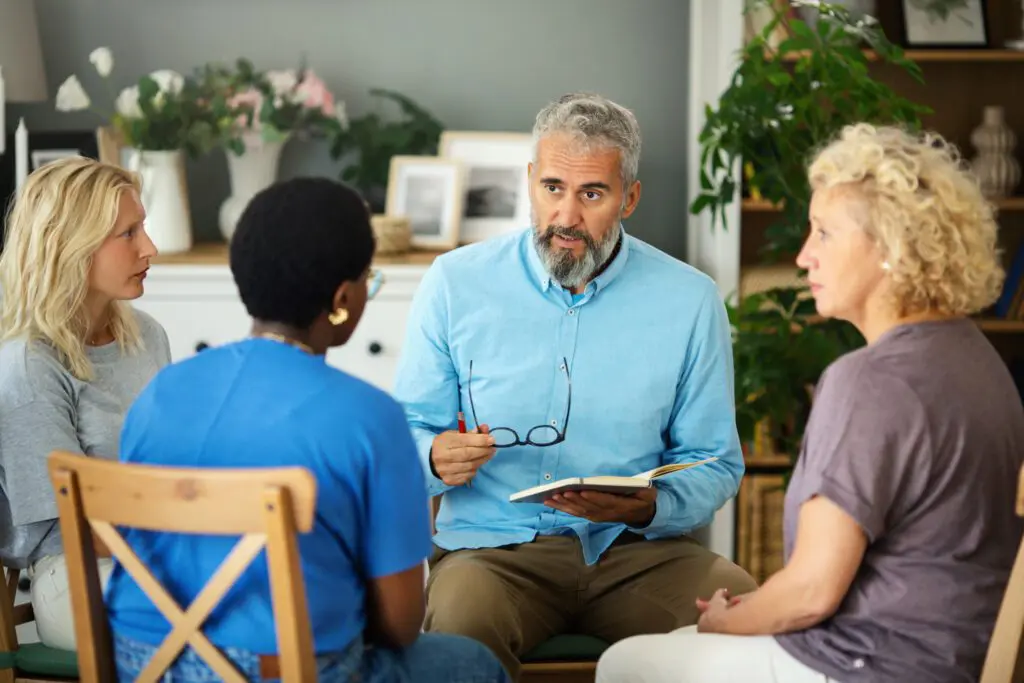 An older man holds a notebook open as he sits in a circle with three woman in a comfortable home-like environment